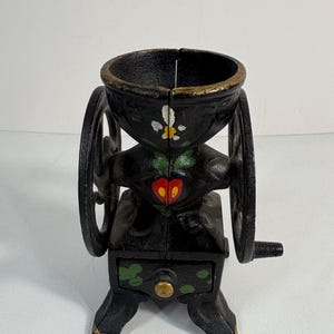 May include: A small, antique black cast iron coffee grinder with a hand crank. The grinder features a decorative design with painted accents of white, green, and red. The grinder has two large wheels and a small drawer.