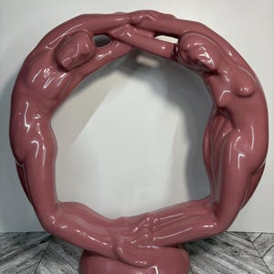May include: A pink ceramic sculpture of two figures intertwined in a circle. The figures are stylised and abstract, with smooth, rounded forms.