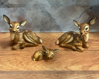 May include: Three ceramic deer figurines in various sizes. The largest deer is brown with white spots and is lying down. The medium-sized deer is also brown with white spots and is lying down. The smallest deer is brown with white spots and is lying on its side.