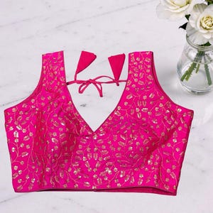 May include: A hot pink, sleeveless blouse with a deep V-neck and intricate gold embroidery. The blouse features a tie closure at the back and is likely made of a smooth, possibly silk-like fabric. The floral embroidery adds a touch of elegance.