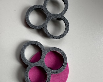 May include: Two dark gray, three-ringed circular frames with a textured surface. One frame is positioned above the other, which is resting on top of a solid, circular, bright pink foam disc. The objects are arranged vertically on a plain surface.