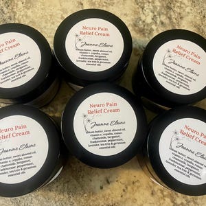 Puede incluir: Seis frascos negros de "Neuro Pain Relief Cream" con etiquetas blancas. Cada etiqueta incluye el nombre "Joanne Elaine" y una lista de ingredientes. Los frascos est&aacute;n dispuestos en un patr&oacute;n circular sobre una superficie clara.