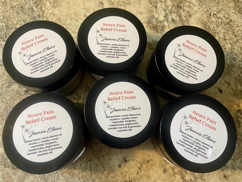 Puede incluir: Seis frascos negros de "Neuro Pain Relief Cream" con etiquetas blancas. Cada etiqueta incluye el nombre "Joanne Elaine" y una lista de ingredientes. Los frascos est&aacute;n dispuestos en un patr&oacute;n circular sobre una superficie clara.