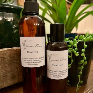 May include: Two amber glass bottles with black pump tops and labels. The labels are white with black text that reads "Joanne Elaine Hanitizer" and lists the ingredients: vodka, aloe, vitamin E, clove, lemon, cinnamon bark, eucalyptus & rosemary essential oils.
