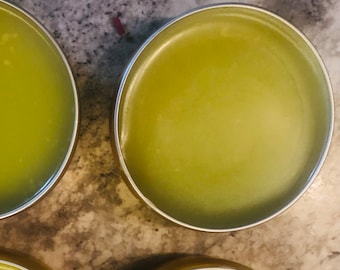 May include: Close-up of several small, round metal tins filled with a light green, creamy substance. The tins are arranged on a speckled, gray surface. The contents of the tins appear smooth and have a slight sheen.