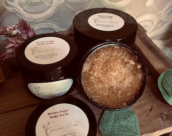 May include: Four dark brown jars of brown sugar body scrub are arranged on a wooden surface. One jar is open, revealing the granular scrub. The labels read "Brown Sugar Body Scrub" and "Joanne Elaine", with ingredient lists.