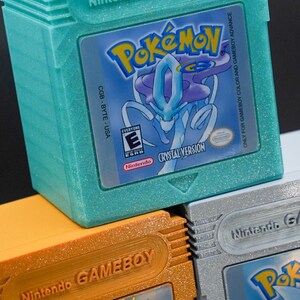 Puede incluir: Tres cartuchos de Game Boy Color apilados uno encima del otro. El cartucho superior es de color turquesa con purpurina y presenta el juego Pokemon Crystal Version. El cartucho del medio es de color naranja con purpurina y presenta el juego Pokemon Gold Version. El cartucho inferior es de color plateado con purpurina y presenta el juego Pokemon Silver Version.