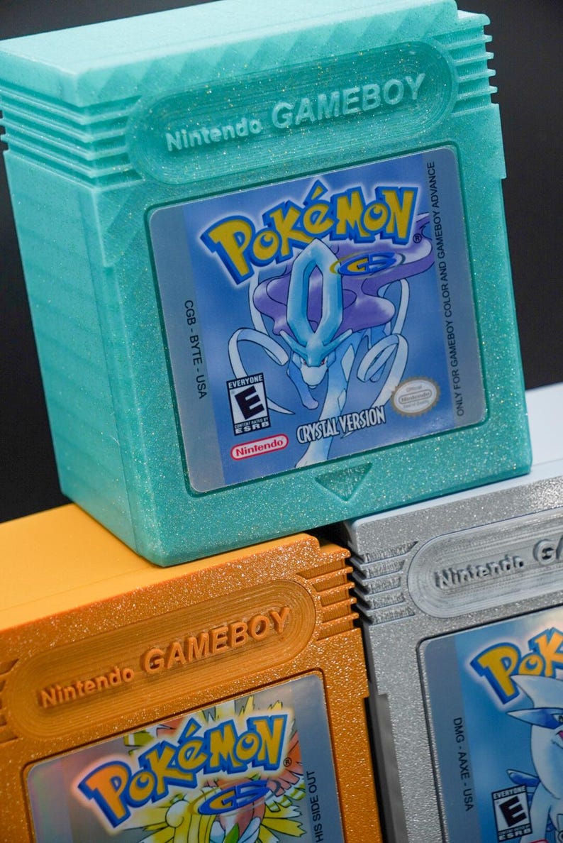 Puede incluir: Tres cartuchos de Game Boy Color apilados uno encima del otro. El cartucho superior es de color turquesa con purpurina y presenta el juego Pokemon Crystal Version. El cartucho del medio es de color naranja con purpurina y presenta el juego Pokemon Gold Version. El cartucho inferior es de color plateado con purpurina y presenta el juego Pokemon Silver Version.