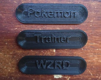 Puede incluir: Tres placas de identificación negras, rectangulares, impresas en 3D con bordes redondeados. La placa superior dice "Pokemon", la placa central dice "Trainer" y la placa inferior dice "WZRD". Las placas de identificación están sobre una superficie de madera.