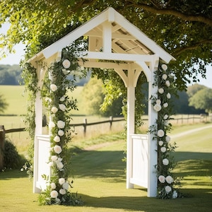 May include: White wooden wedding archway decorated with white flowers and greenery. The archway is standing in a grassy field with a wooden fence in the background.