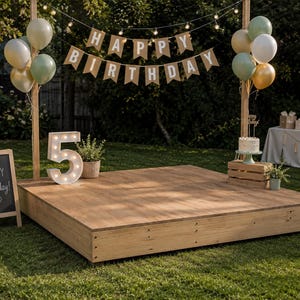 May include: A wooden birthday party stage set outdoors on grass, featuring a Happy Birthday banner, clusters of sage green, white, and gold balloons, a large illuminated number 5, a chalkboard sign with Happy Birthday text, and a cake on a crate.