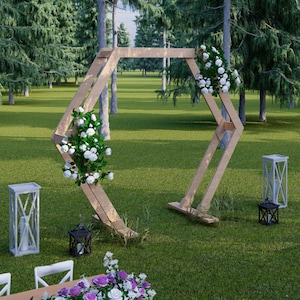 May include: A wooden wedding arch decorated with white flowers and greenery stands on a grassy lawn. Two white lanterns and two black lanterns are placed nearby. A table with white chairs and floral arrangements is in the foreground.