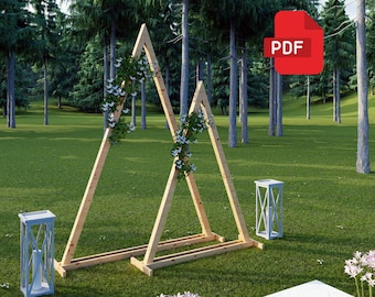 Può includere: Un arco di matrimonio in legno con due telai triangolari decorati con fiori bianchi. L'arco è allestito all'aperto su un prato erboso con alberi sullo sfondo.