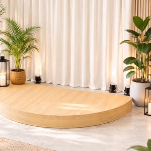 May include: A light wood, semi-circular stage is the focal point, surrounded by potted plants and lit by spotlights and lanterns. The backdrop features white curtains and a wooden panel wall, creating a bright, natural setting.