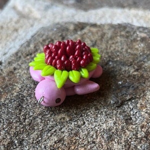 May include: A small pink turtle figurine with a red and green flower on its back. The flower is made of small red beads and green leaves.