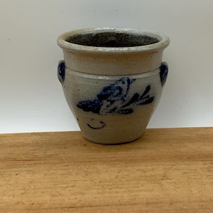 May include: A small, light grey stoneware crock featuring a flared rim and two small side handles. The exterior is decorated with a hand-painted cobalt blue floral motif on the front. The vessel has a tapered base and a smooth, matte finish.