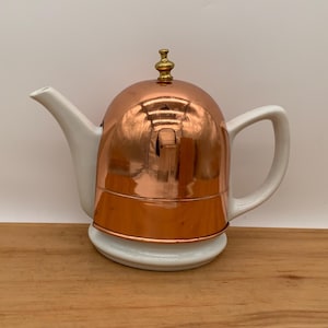 May include: A white and copper teapot with a rounded copper lid and a white spout and handle. The teapot is on a wooden surface against a white background. The copper lid has a small gold-coloured finial.