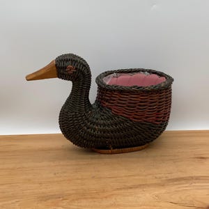 May include: A duck-shaped basket planter with a dark brown woven body and a reddish-brown accent on the lower portion. The duck has a wooden beak and a pink interior. The planter is sitting on a wooden surface.