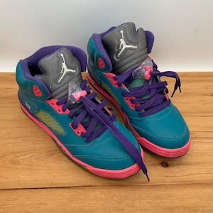 May include: A pair of teal and purple high-top sneakers with pink accents. The sneakers feature purple laces, a gray tongue with a white logo, and a pink sole. The shoes are displayed on a wooden surface.