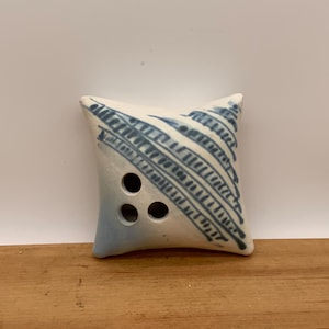 Peut inclure: Petit objet carré en céramique blanche et bleue. Il présente trois trous circulaires et des lignes diagonales. L'objet repose sur une surface en bois devant un fond blanc.