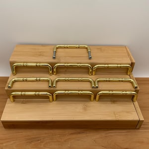 May include: A display of gold-coloured cabinet handles on a wooden stand. The handles are curved with a central bar. The arrangement shows multiple handles, highlighting their design and the quantity available.