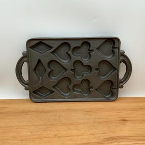 Peut inclure: Un moule en fonte gris foncé pour faire des friandises en forme de cœur, de carreau et de trèfle. La poêle rectangulaire a deux poignées et est conçue avec plusieurs cavités pour la cuisson. Le moule est posé sur une surface en bois.
