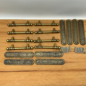 May include: A set of eight brass-toned metal drawer pulls with matching rectangular backplates and a collection of small silver-toned mounting screws. The pulls feature a cylindrical bar design with rounded ends and decorative ring details.