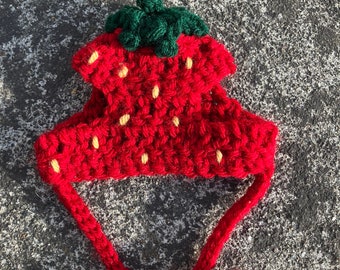 May include: A handmade red crochet strawberry hat with a green stem and yellow seeds. The hat has two long red ties. This whimsical accessory is perfect for a child's costume or a fun photo prop.