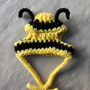 May include: A handmade, yellow and black crocheted bee hat with two black antennae. The hat has a striped design and yellow ties to secure it. This pet accessory is designed to resemble a bee.