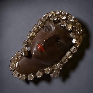 May include: A brooch featuring a dark brown sculpted face with painted red lips, framed by a border of clear, faceted, round rhinestones set in a metallic base. The design depicts a stylized head and shoulders with a glossy finish.