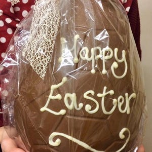May include: A large chocolate Easter egg, wrapped in cellophane, with the words "Happy Easter" piped in white icing. The egg is decorated with a cream-colored bow. A festive treat for the Easter holiday.