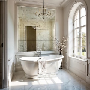 May include: A white freestanding oval bathtub with a chrome faucet and a draped white towel. Behind the tub is a large grid-patterned antique mirror reflecting a chandelier. The room features white marble floors and an arched window.