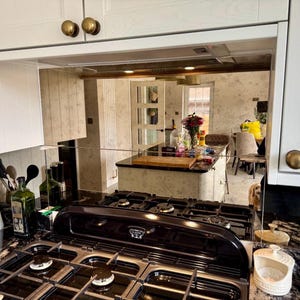 May include: A contemporary kitchen featuring white cabinets with brass handles and a black hob. The hob has multiple burners, and a mirrored splashback reflects the kitchen island and other features.