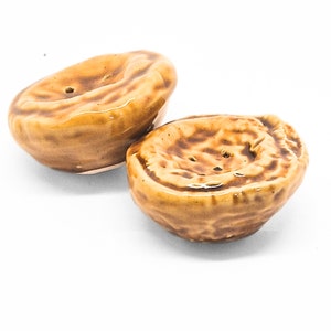 Op de afbeelding: Twee keramische zout- en peperstrooiers in de vorm van gehalveerde walnoten. De strooiers zijn bruin met een geglazuurde afwerking en hebben kleine gaatjes om zout en peper te strooien.