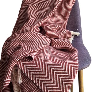 May include: A woven throw blanket featuring a deep red and white herringbone pattern. The fabric displays a consistent zigzag texture throughout the material, and the edges are finished with short, light-colored fringe tassels at the corners.