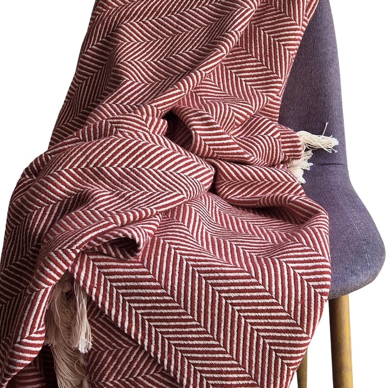 May include: A woven throw blanket featuring a deep red and white herringbone pattern. The fabric displays a consistent zigzag texture throughout the material, and the edges are finished with short, light-colored fringe tassels at the corners.