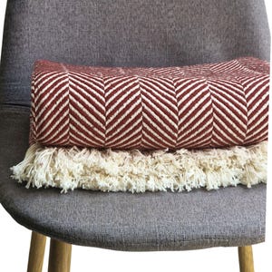 May include: A folded throw blanket featuring a deep red and off-white herringbone pattern. The blanket is finished with a thick, cream-colored fringe along the bottom edge, providing a textured contrast to the woven fabric of the main body.