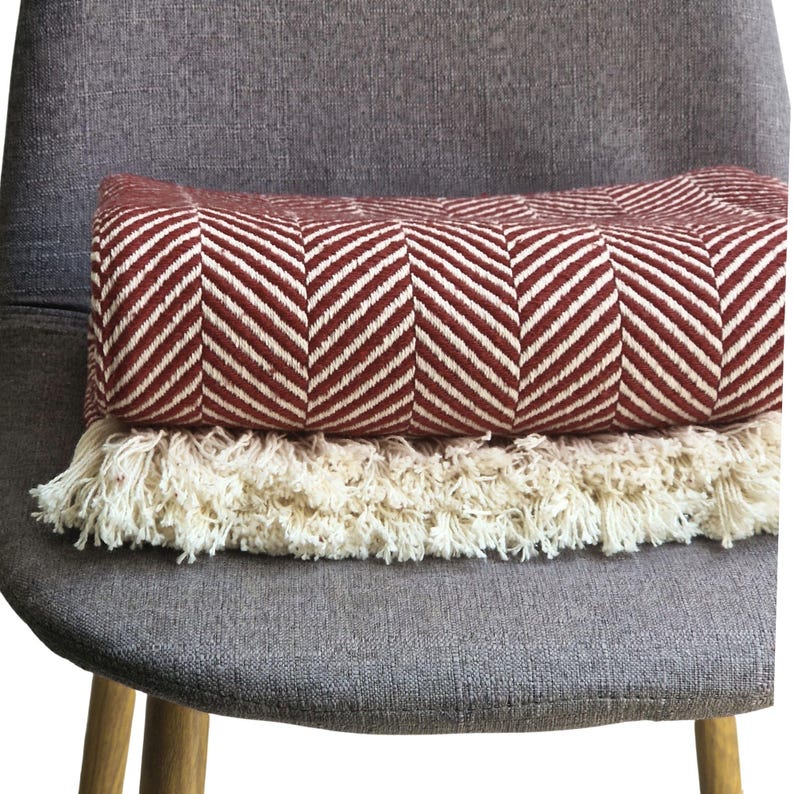 May include: A folded throw blanket featuring a deep red and off-white herringbone pattern. The blanket is finished with a thick, cream-colored fringe along the bottom edge, providing a textured contrast to the woven fabric of the main body.