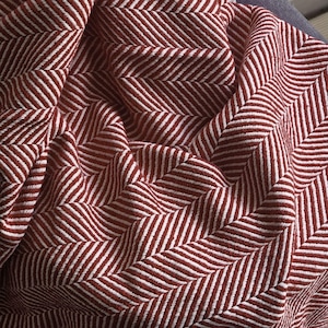 May include: A textile fabric featuring a woven herringbone pattern in deep brick red and crisp white. The material is gathered into soft folds, highlighting the textured weave and the repeating V-shaped geometric design across the surface.