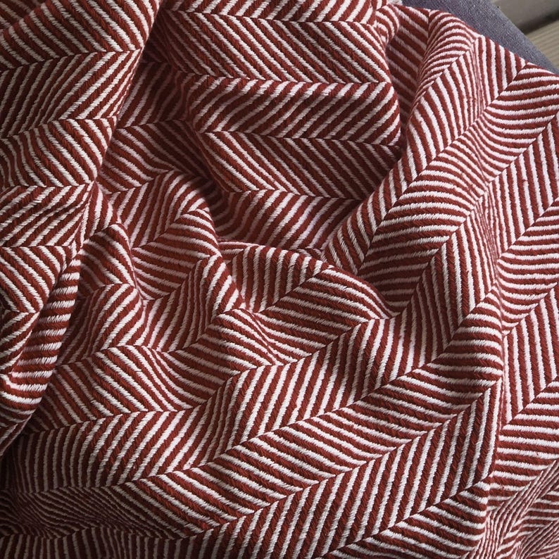 May include: A textile fabric featuring a woven herringbone pattern in deep brick red and crisp white. The material is gathered into soft folds, highlighting the textured weave and the repeating V-shaped geometric design across the surface.