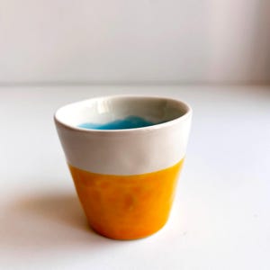 Op de afbeelding: Een klein, wit keramiek kopje met een blauwe binnenkant en een gele buitenkant. Het kopje is geglazuurd en heeft een gladde afwerking.