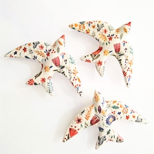 Pode incluir: Três ornamentos de parede em forma de pássaro em cerâmica de cor creme, cada um com um padrão floral pintado à mão em tons de vermelho, laranja, amarelo, azul e verde. Os pássaros são retratados em posição de voo com asas abertas e caudas.