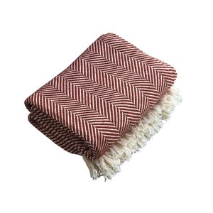 May include: A folded throw blanket featuring a deep red and white herringbone pattern. The textile has a woven texture and is finished with long, cream-colored fringe tassels along the bottom edge, creating a distinct contrast against the pattern.