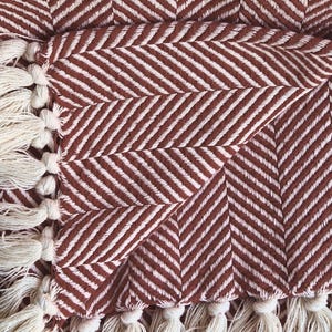 May include: A woven throw blanket featuring a rust-colored and cream herringbone pattern. The fabric displays a distinct V-shaped weave texture throughout. The edges of the blanket are finished with a row of knotted cream-colored fringe tassels.