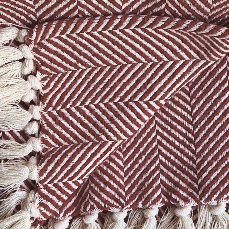 May include: A woven throw blanket featuring a rust-colored and cream herringbone pattern. The fabric displays a distinct V-shaped weave texture throughout. The edges of the blanket are finished with a row of knotted cream-colored fringe tassels.