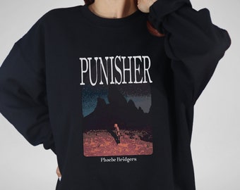 Op de afbeelding: Zwart sweatshirt met een witte grafische print van de tekst "PUNISHER" en een landschapsscène met een persoon op de voorgrond. De tekst "Phoebe Bridgers" is onder de grafische print gedrukt.
