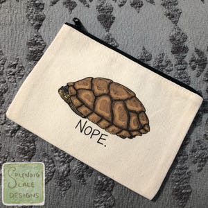 May include: Cream-colored canvas pouch with a black zipper and a turtle shell illustration. The turtle shell is brown with darker brown shading. The word "NOPE" is printed in black below the turtle shell.