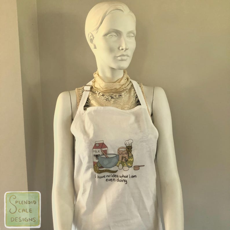 Op de afbeelding: Witte schort met een kleurrijke illustratie van bakbenodigdheden en een mengkom. De schort heeft de tekst "i have no idea what i am even doing" onder de illustratie. De schort wordt getoond op een mannequin.
