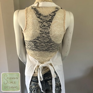 Op de afbeelding: Een cr&egrave;mekleurig kanten vest met een zwart kanten achterpaneel wordt gedragen over een wit overhemd met kraag en een spijkerbroek. Een witte schort is om de taille geknoopt. Het vest staat op een mannequin.