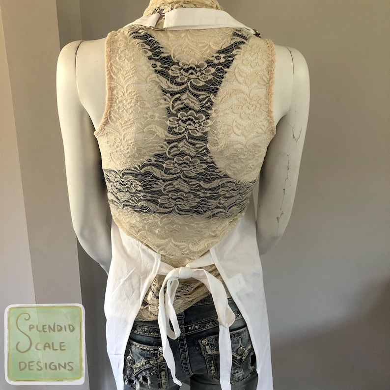 Op de afbeelding: Een cr&egrave;mekleurig kanten vest met een zwart kanten achterpaneel wordt gedragen over een wit overhemd met kraag en een spijkerbroek. Een witte schort is om de taille geknoopt. Het vest staat op een mannequin.